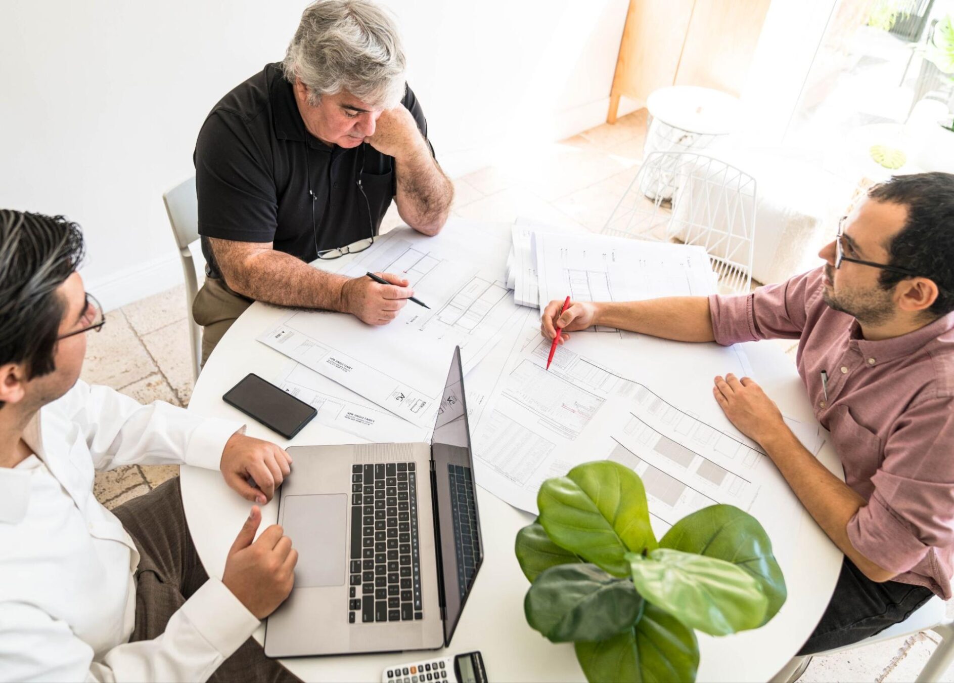 A round white table is covered in papers, charts, and a laptop as three investors work to understand the difference between the Investing vs. Real Estate Development question and it's impact for ROI.