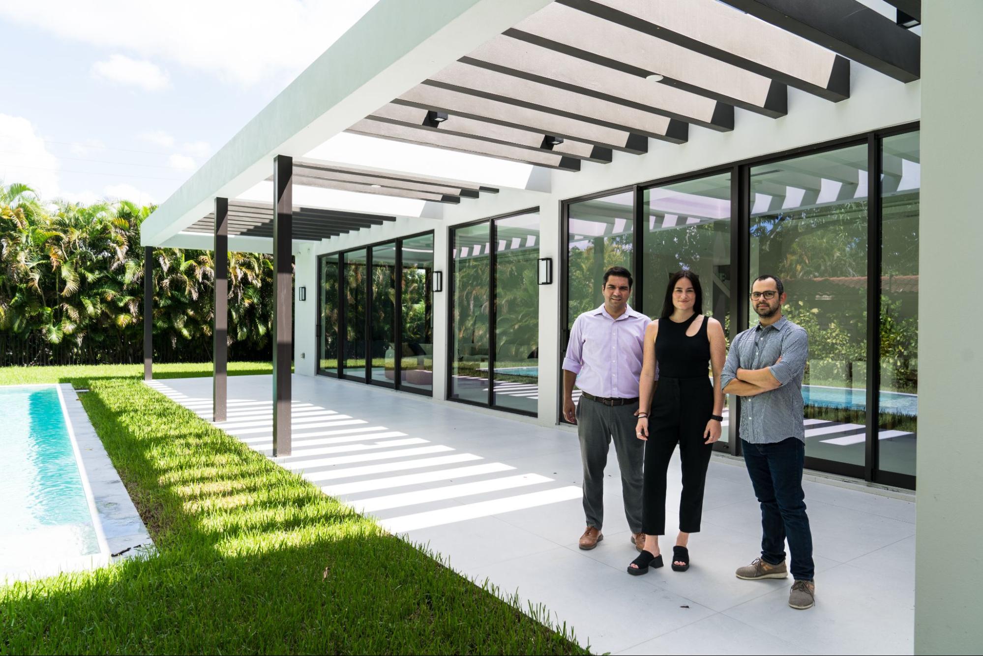 Three well-dressed people stand under a covered porch of a luxurious modern home in Miami.